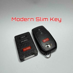 May include: Two black car key fobs with red accents and the text "Modern Slim Key." One fob has buttons for lock, unlock, and alarm. The other has buttons for lock, unlock, and a red "HOLD" button.