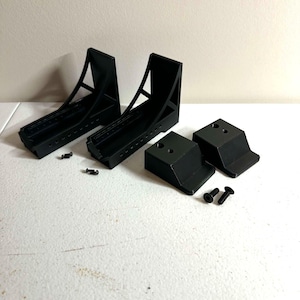 May include: Two black L-shaped brackets with open-frame support structures, two smaller black wedge-shaped mounting blocks, and four small black screws. The components are made of a matte black material and feature pre-drilled holes for assembly.