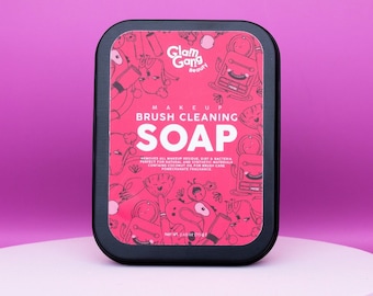 May include: A black tin container with a pink label featuring a cartoon design. The label reads "Glam Gang Beauty" and "Makeup Brush Cleaning Soap". The text on the label also states "Removes all makeup residue, dirt & bacteria. Perfect for natural and synthetic materials. Contains coconut oil for brush care. Pomegranate fragrance."