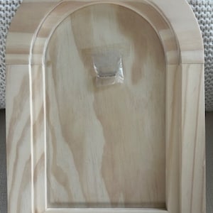 May include: A light-colored, unfinished wooden arch-shaped frame. The frame has a rectangular base with an arched top, featuring a recessed interior. The wood grain is visible throughout the piece. A small plastic packet is attached to the back.