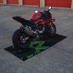 May include: A red and black sport motorcycle parked on a black and green graphic mat. The motorcycle has a sleek design with visible exhaust pipes and mirrors. The background features red storage doors.
