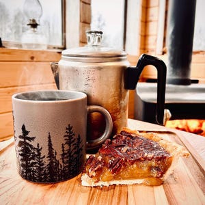 May include: A grey ceramic mug with a black silhouette of pine trees, a metallic percolator coffee pot with a black handle, and a slice of pecan pie on a wooden board. The mug contains a steaming hot beverage, and the pie has a golden crust.