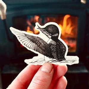 May include: A black and white sticker featuring a detailed illustration of a loon with outstretched wings. The bird is depicted in a detailed, artistic style, with intricate feather patterns. The sticker is held in front of a blurred background of a fireplace.