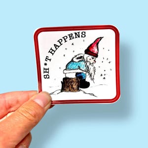 May include: A square sticker with a red border featuring a cartoon gnome sitting on a tree stump in a snowy scene. The gnome wears a red hat and blue coat. The text "SH*T HAPPENS" is printed around the edge of the sticker.