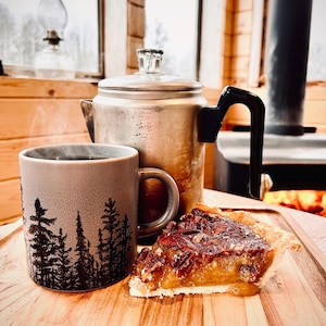 May include: A rustic scene featuring a coffee mug with a forest silhouette, a vintage metal coffee pot, and a slice of pecan pie on a wooden tray. The mug contains a hot beverage. The pie has a golden filling and pecan topping.