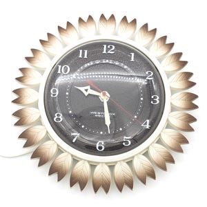 Può includere: Un orologio bianco e marrone a forma di girasole con lancette nere e un quadrante nero. Il quadrante dell'orologio ha il testo "WESTOX ELECTRIC" e i numeri da 1 a 12.