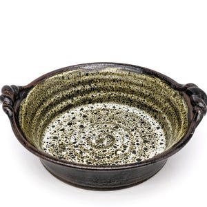 Puede incluir: Un plato para hornear redondo de cerámica con dos asas. El plato tiene un esmalte moteado en tonos de verde, marrón y blanco.