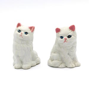 Könnte beinhalten: Zwei weiße Keramik-Katzenfiguren mit rosa Ohren und blauen Augen. Die Katzen sitzen mit ihren Pfoten unter sich.