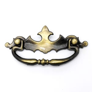 以下が含まれることがあります： 装飾的な装飾が施された、金色と黒の金属製のキャビネットプル。プルには湾曲したハンドルと装飾的なトップがあります。