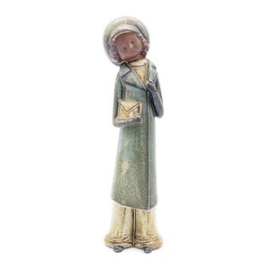Może przedstawiać: Ceramiczna figurka przedstawiająca osobę w zielonym płaszczu i brązowym kapeluszu, trzymającą brązową kopertę. Figurka ma brązowe włosy i brązowe oczy.