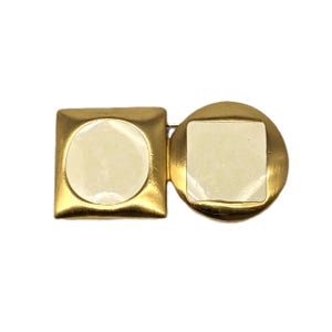 Puede incluir: Broche dorado con un cuadrado y un círculo, cada uno con una incrustación crema, circular y cuadrada. El broche tiene una estética vintage y probablemente está hecho de metal.