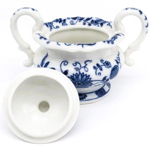 Pu&ograve; includere: Una zuccheriera in porcellana bianca con un motivo floreale blu e bianco. La ciotola ha due manici e un coperchio. Il coperchio &egrave; aperto e l'interno della ciotola &egrave; visibile.
