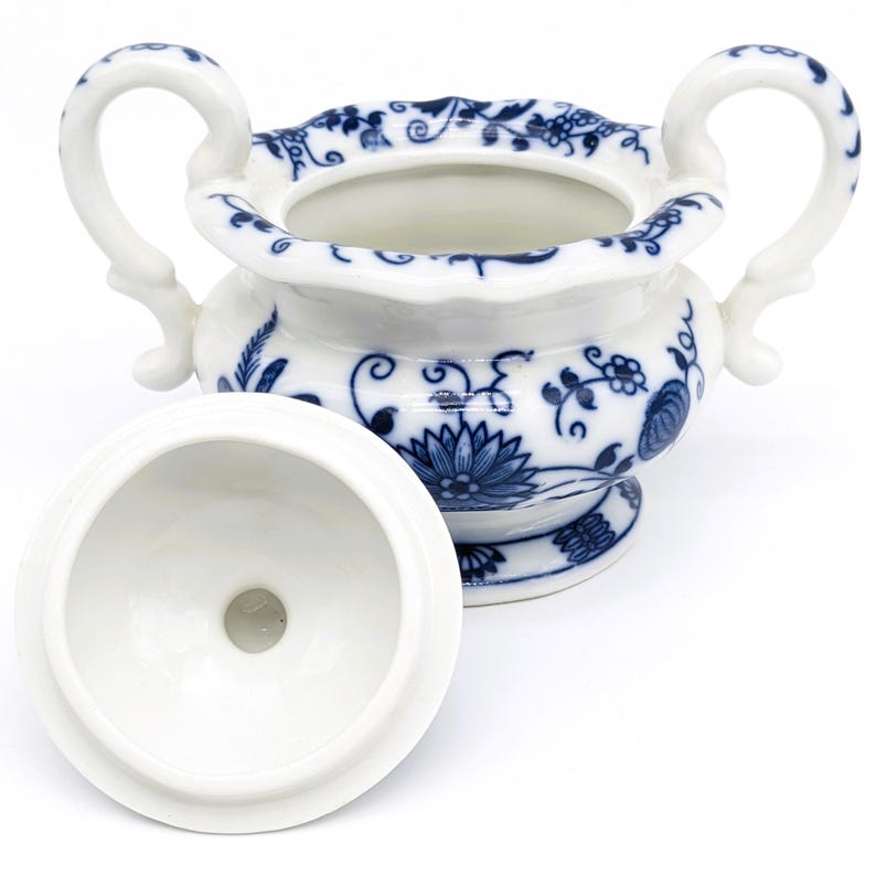 Pu&ograve; includere: Una zuccheriera in porcellana bianca con un motivo floreale blu e bianco. La ciotola ha due manici e un coperchio. Il coperchio &egrave; aperto e l'interno della ciotola &egrave; visibile.