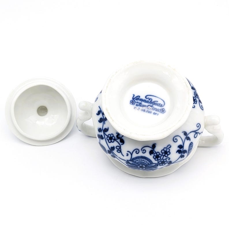 Pu&ograve; includere: Una zuccheriera in porcellana bianca con un motivo floreale blu e bianco. La zucchieriera ha un coperchio e due manici. Sul fondo della zucchieriera &egrave; presente il testo "Vienna House Fine China" in blu.
