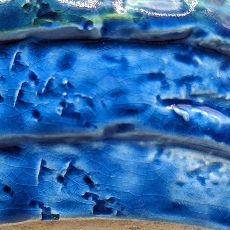 Op de afbeelding: Close-up van een blauw keramisch oppervlak met een gekrakelde glazuur. Het oppervlak heeft een textuur en een onregelmatige uitstraling.