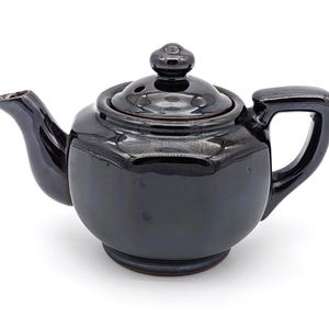Può includere: Una teiera in ceramica nera con un coperchio e un beccuccio. La teiera ha una finitura lucida e un corpo arrotondato.