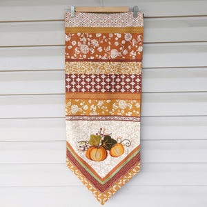 Peut inclure: Un chemin de table long et rectangulaire avec un motif patchwork en différentes nuances d'orange, de brun et de blanc. Le chemin de table a une extrémité pointue et présente un motif brodé de deux citrouilles avec des feuilles et des vignes.