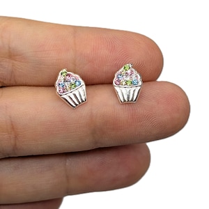 Puede incluir: Un par de pendientes de botón de cupcake plateados. Cada pendiente presenta un diseño de cupcake con una base plateada y detalles de piedras preciosas de colores en la parte superior. Las piedras preciosas incluyen rosa, azul, verde y amarillo.