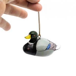 Puede incluir: Un adorno de madera en forma de pato con cabeza verde, pico amarillo y cuerpo negro y gris. El pato está pintado con detalles azules y rosas en las alas. El adorno tiene una cuerda dorada para colgar.