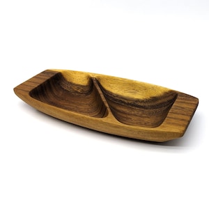 Puede incluir: Una fuente rectangular de madera con dos compartimentos. La fuente está hecha de madera marrón clara y oscura, con un acabado liso y pulido. Los compartimentos tienen forma de cuenco y están diseñados para contener aperitivos o salsas.