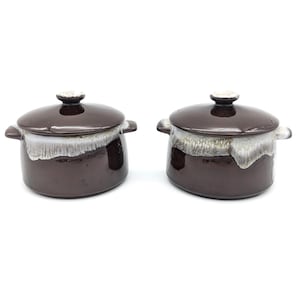 May include: Two brown ceramic casserole dishes with lids. Each dish has a white and tan glaze around the rim and handles. The lids have a small, round knob on top. The dishes are on a white background.