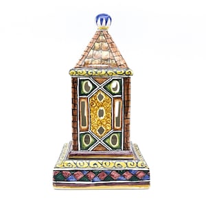 Peut inclure: Une lanterne décorative en céramique avec un design de tour. La lanterne présente un motif de briques marron, un toit en tuiles et des motifs géométriques colorés. Le dessus est orné d'un fleuron bleu et la base a une bordure décorative.