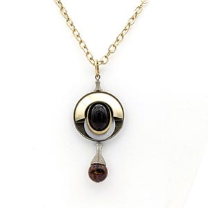 May include: A gold chain necklace with a pendant featuring a black oval gemstone surrounded by a silver and gold circular design. The pendant has a small brown bead hanging from the bottom.