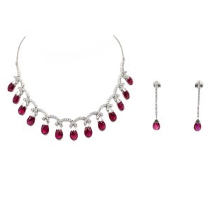 May include: A silver necklace and matching earrings set. The necklace features a series of teardrop-shaped, deep red gemstones suspended from a delicate, curved chain. The earrings have a single teardrop gemstone. The set is displayed against a white background.