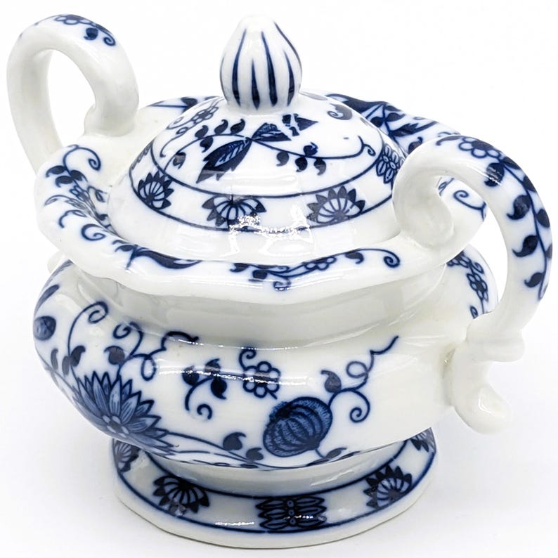 Pu&ograve; includere: Una zuccheriera in porcellana bianca con un motivo floreale blu e bianco. La ciotola ha due manici e un coperchio con un pomo in cima.
