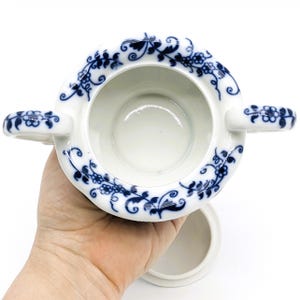 Pu&ograve; includere: Una zuccheriera in porcellana bianca con un motivo floreale blu e bianco. La ciotola ha due manici e un coperchio.