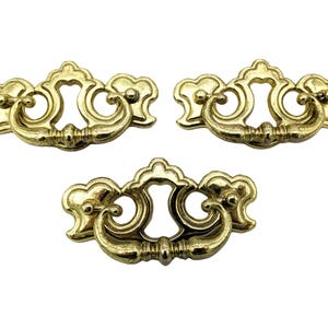 May include: Three ornate gold-toned cabinet drawer pulls with a decorative scroll design.