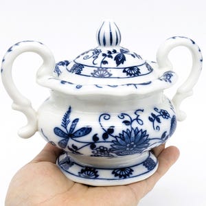 Pu&ograve; includere: Una zuccheriera in porcellana bianca con un motivo floreale blu e bianco. La ciotola ha un coperchio e due manici.