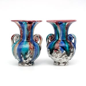 Peut inclure: Deux vases en céramique avec un motif abstrait coloré. Les vases ont un bord évasé, deux anses et une base texturée. La palette de couleurs comprend des nuances de bleu, de turquoise, de rouge et de blanc. Les vases sont sur fond blanc.