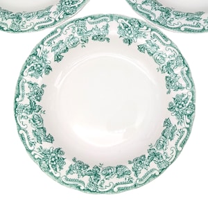 May include: White ceramic plates with a green floral pattern around the rim. The design features roses and other flowers with a scalloped edge. The plates are arranged on a white background.