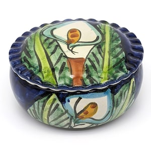 May include: A round ceramic trinket box with a lid, featuring a hand-painted design of white calla lilies with green leaves on a blue background.