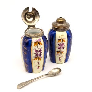 Könnte beinhalten: Ein Paar antike Salz- und Pfefferstreuer aus Keramik mit silberfarbenen Deckeln und einem kleinen Löffel. Die Streuer sind weiß mit blauen vertikalen Streifen, goldenen Akzenten und floralen Designs. Ein Streuer hat einen offenen Deckel.