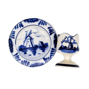 Könnte beinhalten: Ein weiß-blaues Keramik-Teller- und Eierbecher-Set. Der Teller zeigt eine Windmühlenszene mit blauen Akzenten. Der Eierbecher hat ein Windmühlendesign und das Wort "Holland". Das Set ist wahrscheinlich Vintage.