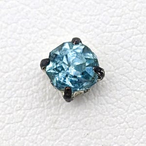May include: A small, round, light blue gemstone with a silver setting and four black prongs.