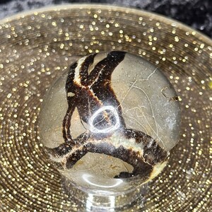 Op de afbeelding: Een gepolijste, ronde, bruine en witte agaatbol met een draaiend patroon. De bol ligt op een gouden glitteroppervlak.