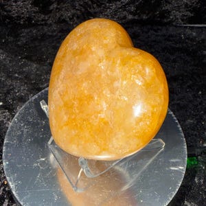 May include: A polished, golden-orange heart-shaped crystal. The gemstone sits on a clear acrylic stand, reflecting light. The heart is smooth and has a natural, textured appearance, perfect for home decor or as a unique gift.