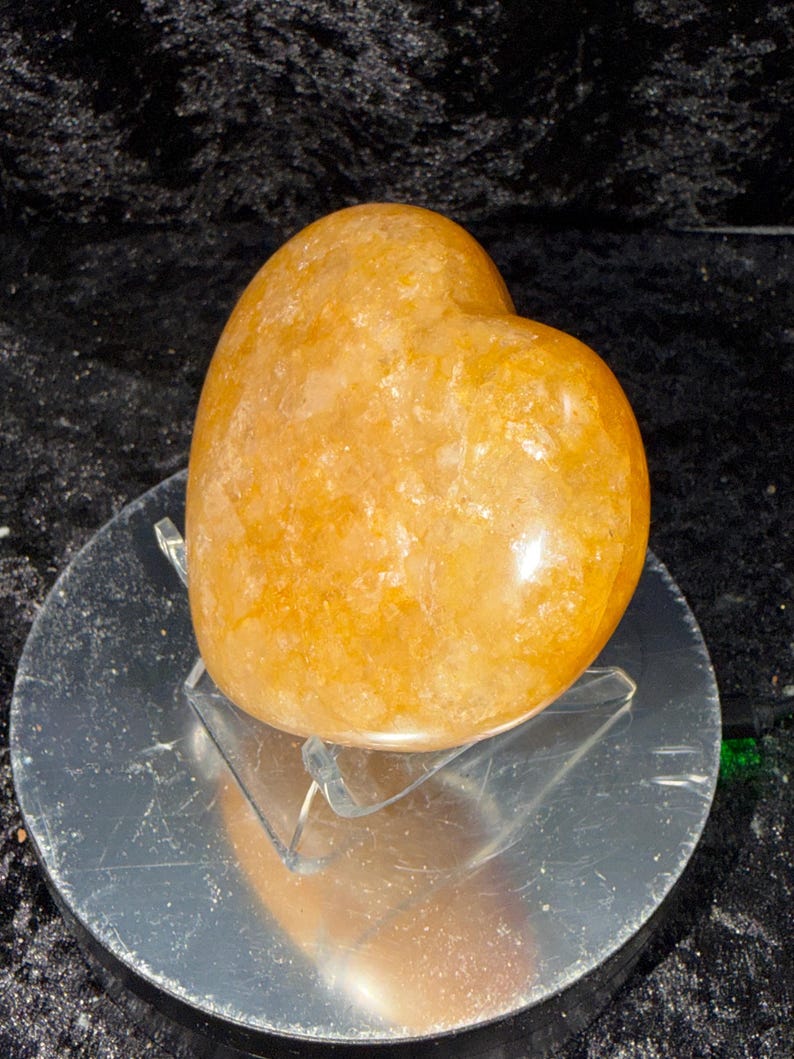 May include: A polished, golden-orange heart-shaped crystal. The gemstone sits on a clear acrylic stand, reflecting light. The heart is smooth and has a natural, textured appearance, perfect for home decor or as a unique gift.