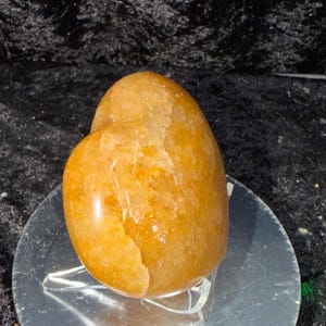 May include: A polished, golden-orange heart-shaped stone. The stone has a smooth, glossy surface with natural variations in color and texture. It sits on a clear, plastic stand against a dark background.