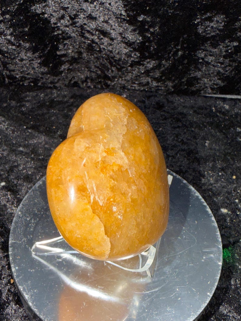 May include: A polished, golden-orange heart-shaped stone. The stone has a smooth, glossy surface with natural variations in color and texture. It sits on a clear, plastic stand against a dark background.