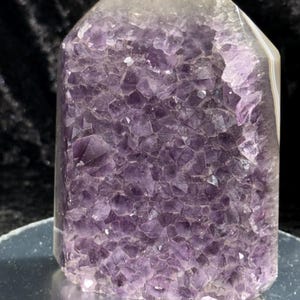 May include: A polished amethyst crystal geode with a flat top and a rectangular shape. The crystal is a deep purple color with a crystalline structure. The geode is sitting on a reflective surface.