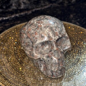 May include: A detailed, carved skull figurine in shades of gray, black, and red granite. The skull is resting on a gold-colored, textured plate. The background is a dark, textured fabric.