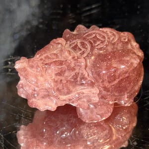 May include: A translucent pink strawberry quartz carving of a mythical creature, likely a Pixiu, featuring intricate details including scales and circular coin motifs along its back. The gemstone has a sparkling, crystalline texture throughout.