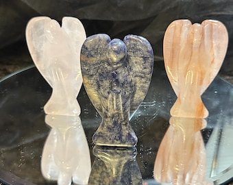 May include: Three carved angel figurines in shades of pink, blue, and peach are displayed on a reflective surface. The angels have detailed wings and are made of polished stone. The background is a dark, textured fabric.