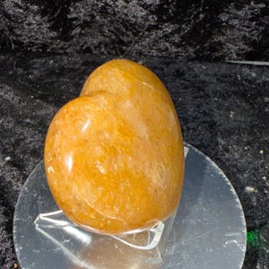 May include: A polished, golden-orange gemstone, possibly a quartz, with a smooth, rounded shape. The stone is displayed on a clear acrylic stand atop a reflective silver surface, set against a dark, textured background.