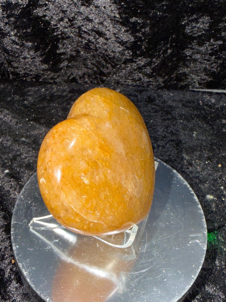 May include: A polished, golden-orange gemstone, possibly a quartz, with a smooth, rounded shape. The stone is displayed on a clear acrylic stand atop a reflective silver surface, set against a dark, textured background.