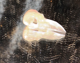 May include: A small, translucent, pale pink carved stone figurine shaped like a bird skull. The object features a pointed beak, a hollow eye socket, and a smooth, polished surface that reflects light, showing a clear silhouette of the avian cranium.